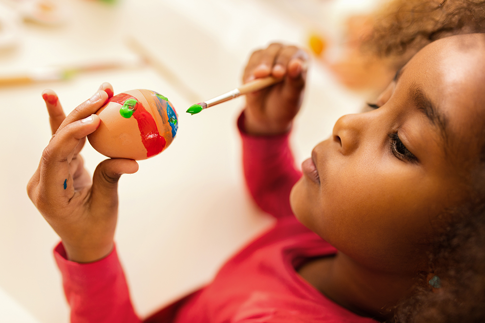 Close up of African American little girl coloring Easter egg with paintbrush.